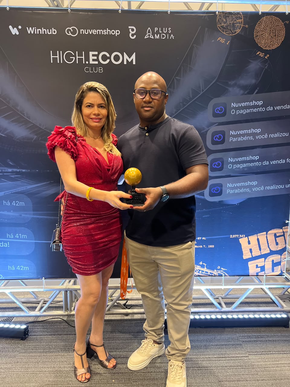 Fotografia completa de Clébão acompanhado no evento High.Ecom, segurando o troféu dourado
