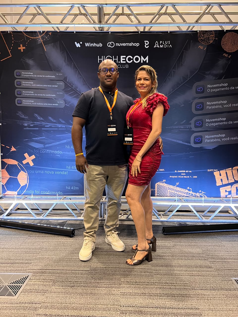 Foto completa de Clébão acompanhado no evento High.Ecom, com os dois de corpo inteiro e o cenário do evento